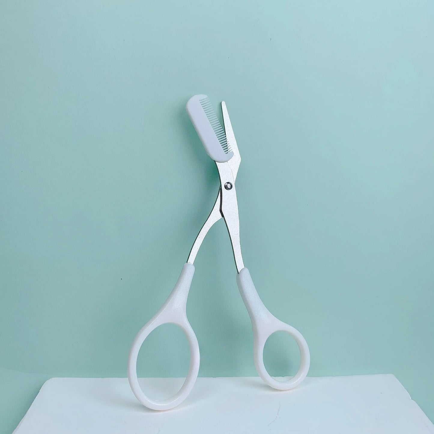 Eyebrow Trimming Scissors with Built-in Comb resting on a light blue background, showcasing its grooming design.